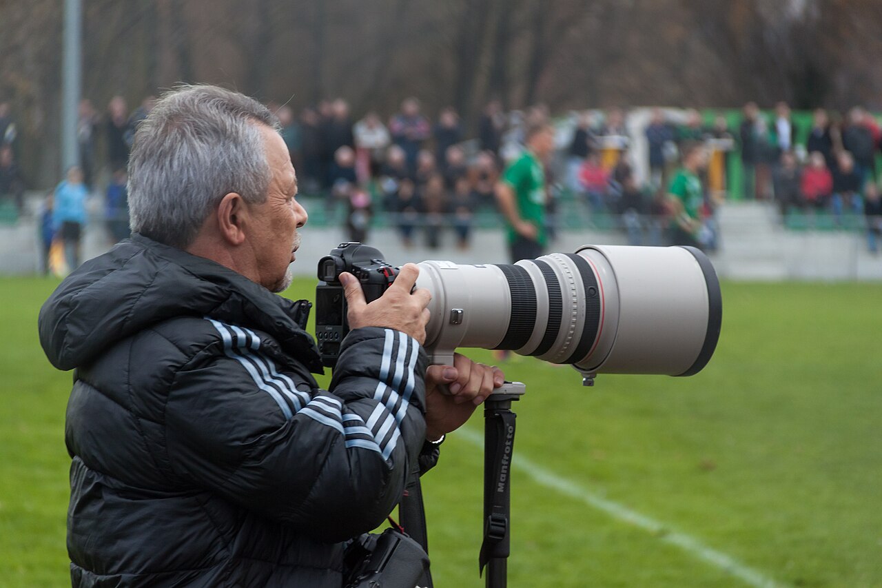 Un photographe accroupi au bord d’un terrain de football, un long téléobjectif à la main